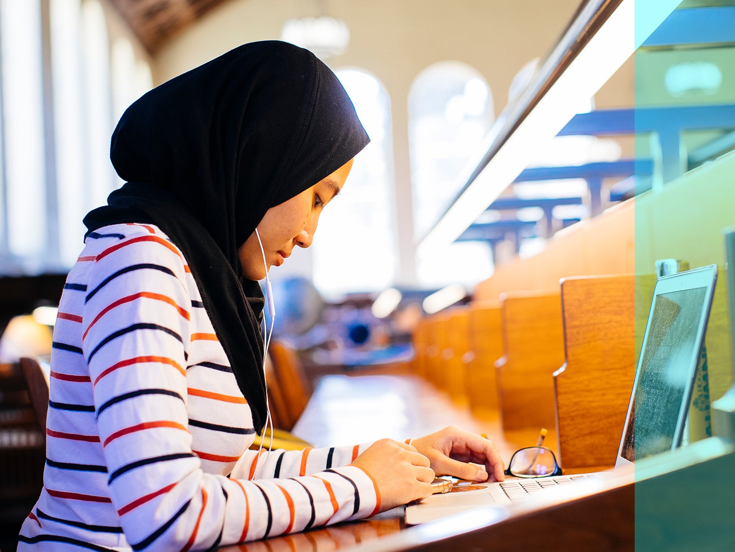 A woman wearing a headscarf does research on a computer in the library.