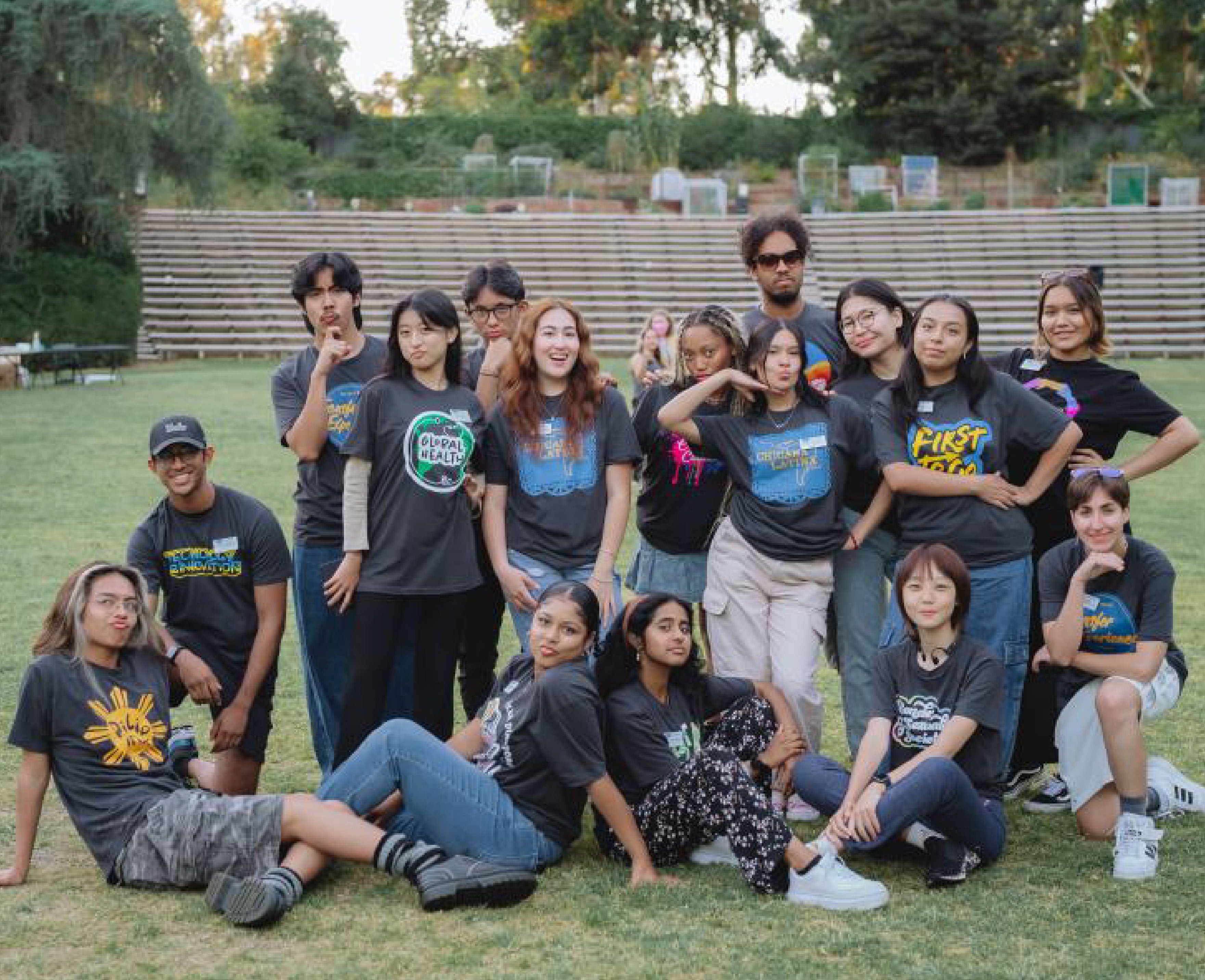 A group photo taken outdoors of diverse students, all wearing various graphic T-shirts, standing and sitting together on a grassy field. The students are smiling and posing playfully, creating a sense of camaraderie.
